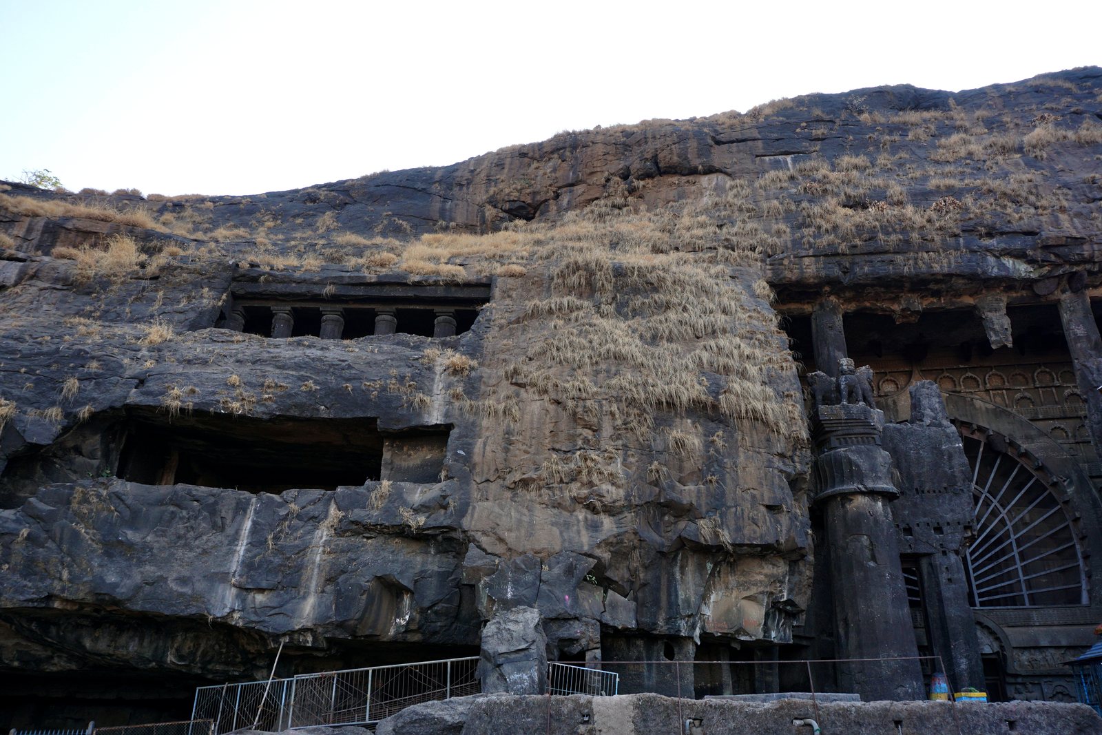 Photograph of the Karla Caves hillside showing rock-cut cave openings, the chaitya hall entrance and a free-standing carved pillar.